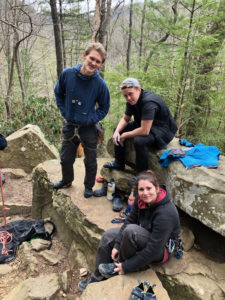 Samantha War celebrates the completion of her most difficult climb to date with two fellow climbers, Nate Valentine (left) and Eric Snoek. They are sitting on a rock face in the woods, surrounded by their climbing gear.