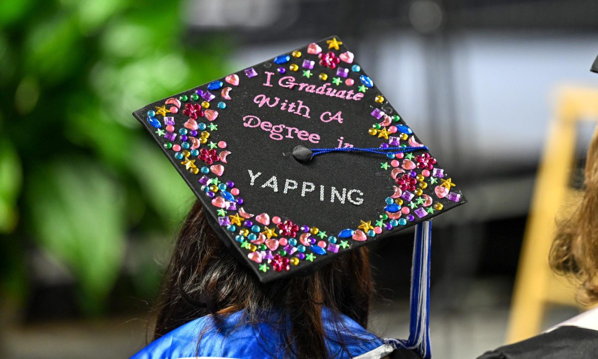 A graduate stands up before walking to the stage with a mortarboard decorated with the phrase "I graduated with a degree in yapping" during spring commencement inside the Murphy Center on May 9, 2026.
