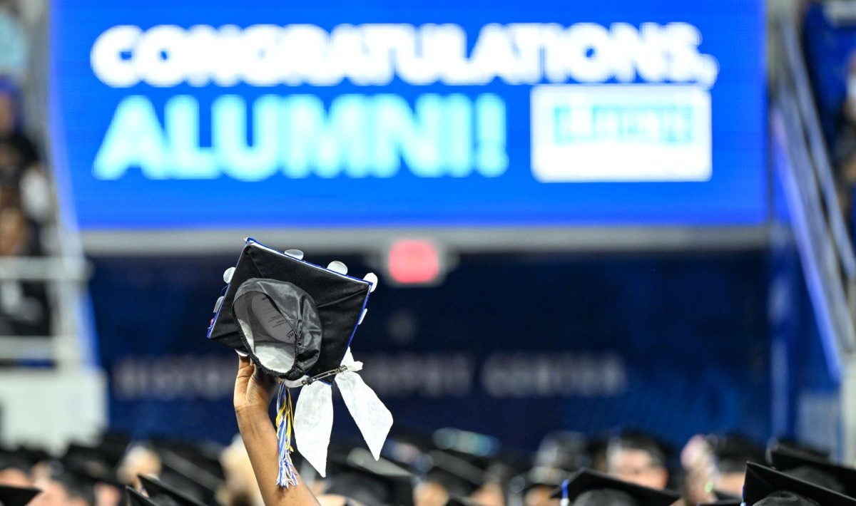 A graduate lifts up their cap in front of a "congratulations alumni" sign during spring commencement inside the Murphy Center on May 9, 2026.